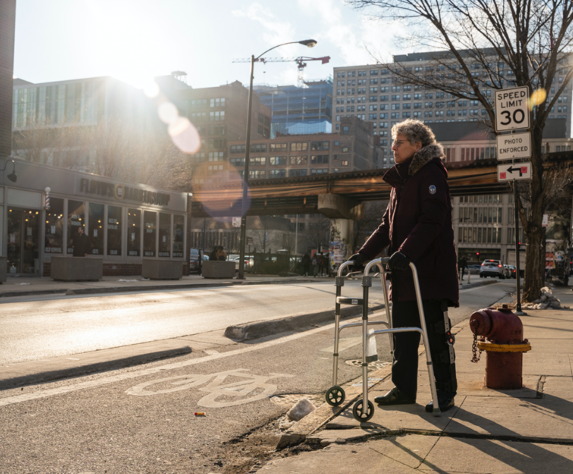 Con Buckley, part-time faculty member in the Humanities, History and Social Sciences Department, stands with her leg brace and walker near the concrete-protected bike lane that she tripped over, shattering her patella.