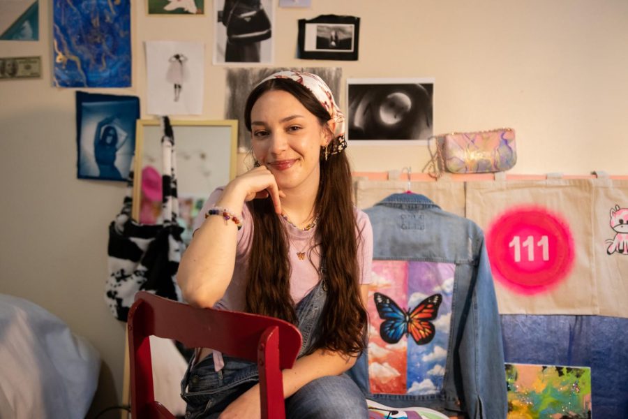 Judith Grace Hoffman, a junior photography major, sits in front of a display of her designs, including a denim jacket with a butterfly and a cow print bag.