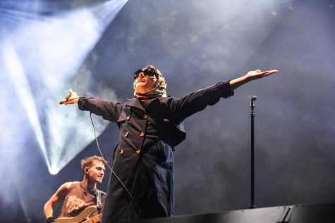 Gerard Way of My Chemical Romance, dressed in a coat and a headwrap, performs energetically on stage as the crowd screams in excitement. 