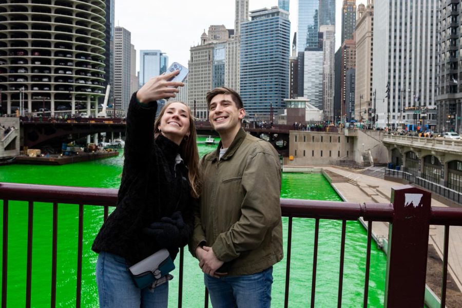 Smiling for a selfie, two people pose, using the brightly green dyed river and Chicago Riverwalk skyline as their backdrop. The river was dyed at 10 a.m. by the Chicago Plumbers Local Union 130.