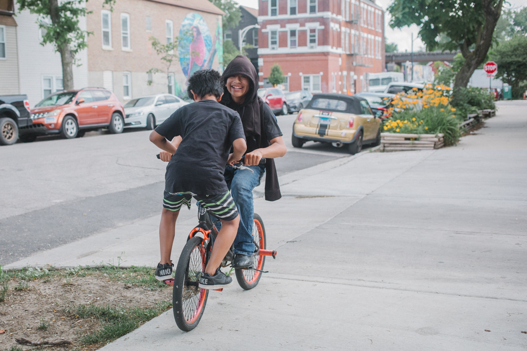 Two young children on a bike, with one sitting on the handlebars, carefully make their way down Wolcott Ave. in Pilsen, Chicago, on Thursday, June 26, 2025