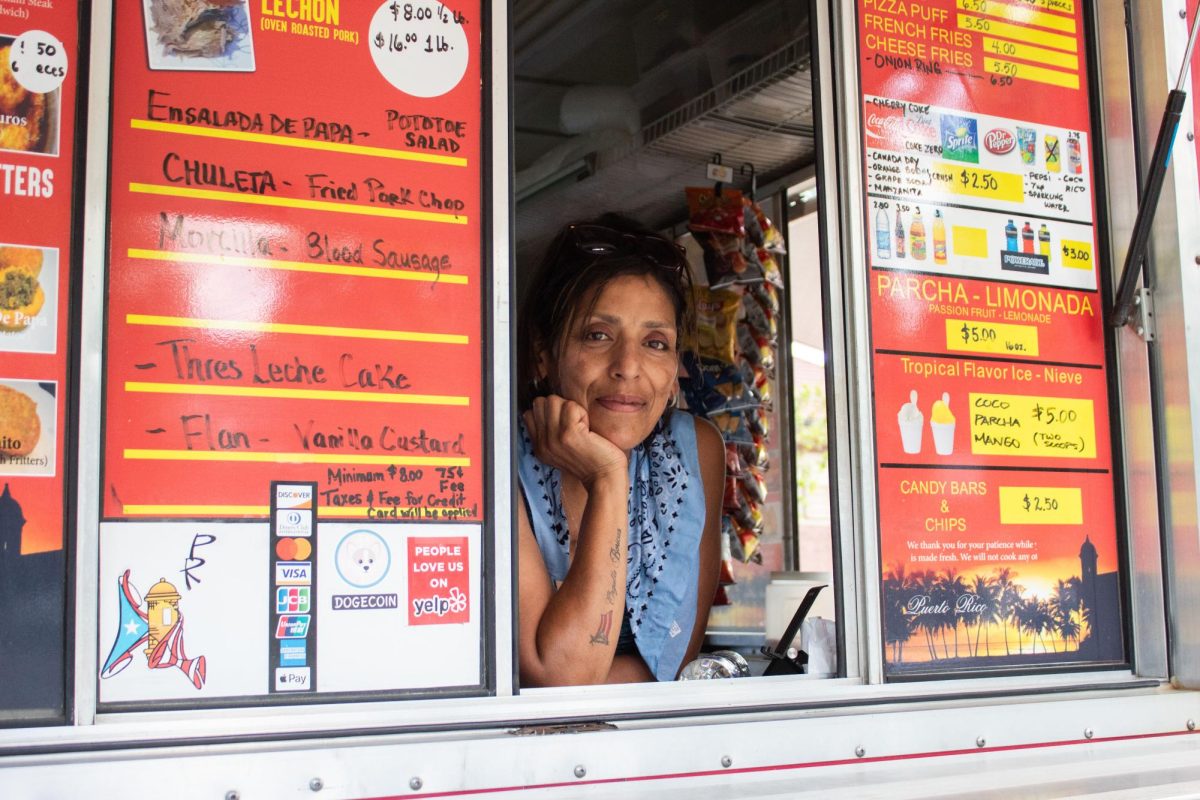 María Cáceres leans out of the service window of her food truck, Snix Snacks, at Independence Park in Chicago's Irving Park neighborhood on Thursday, July 2, 2026. She has sold Puerto Rican dishes there for 13 years. This photo is part of the Columbia Chronicle’s “77 Community Areas” project, documenting Chicago ahead of America’s 250th birthday.