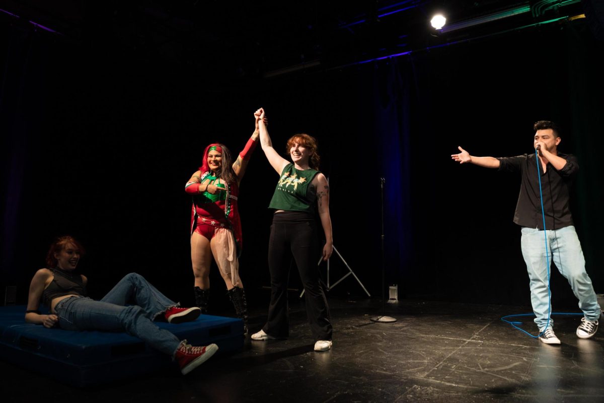 La leyenda de lucha libre profesional Paloma "La Sriracha Muchacha" Vargas, a la izquierda, y la estudiante Abigail Ehrenberg, a su lado, alzan sus manos tras una demostración en el escenario, mientras Shelbee Taylor, abajo a la izquierda, y un presentador en el micrófono, observan durante el taller de lucha libre en Columbia College Chicago, el martes 13 de octubre de 2026.